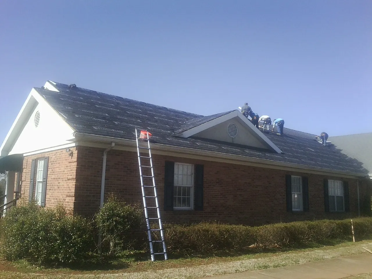 Crew tearing shingles off a brick ranch roof with a ladder set — Metal Roof Repair work by our crew in Larwill