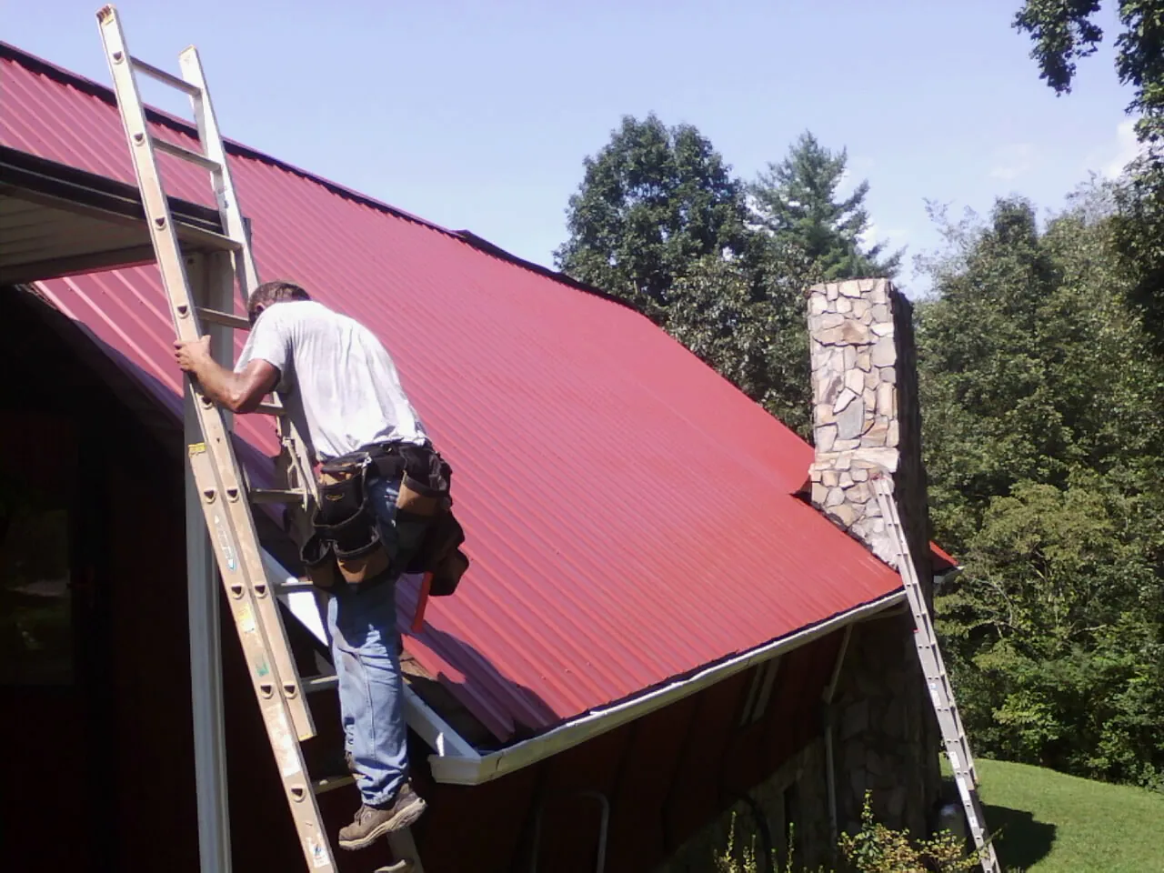 Roofer on a ladder beside a red metal roof and stone chimney — Soffit & Fascia Repair by Roofing Brothers in Larwill